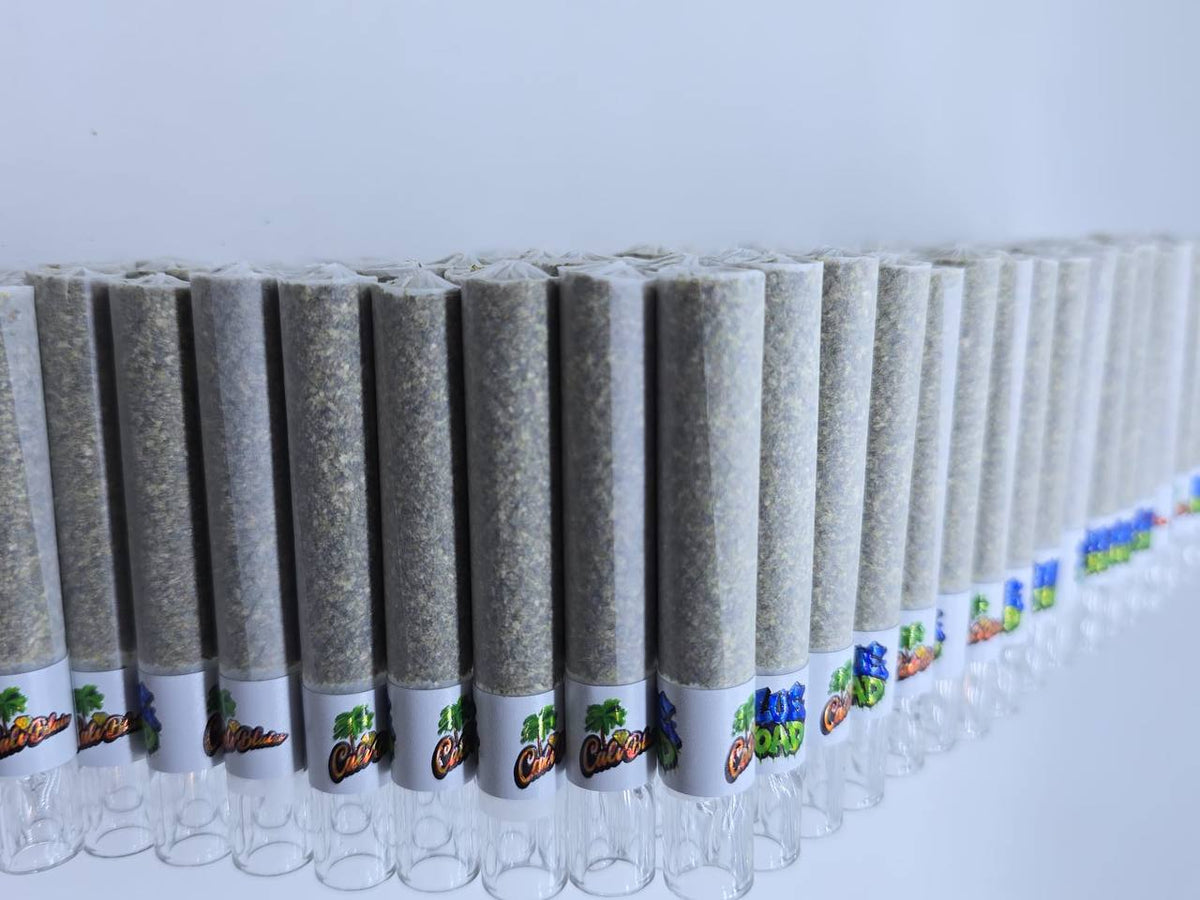A diagonal row of rolling paper joints filled with a light gray-green herbal blend, lined with white paper and decorated with colorful cartoon-style labels, set against a plain white background.